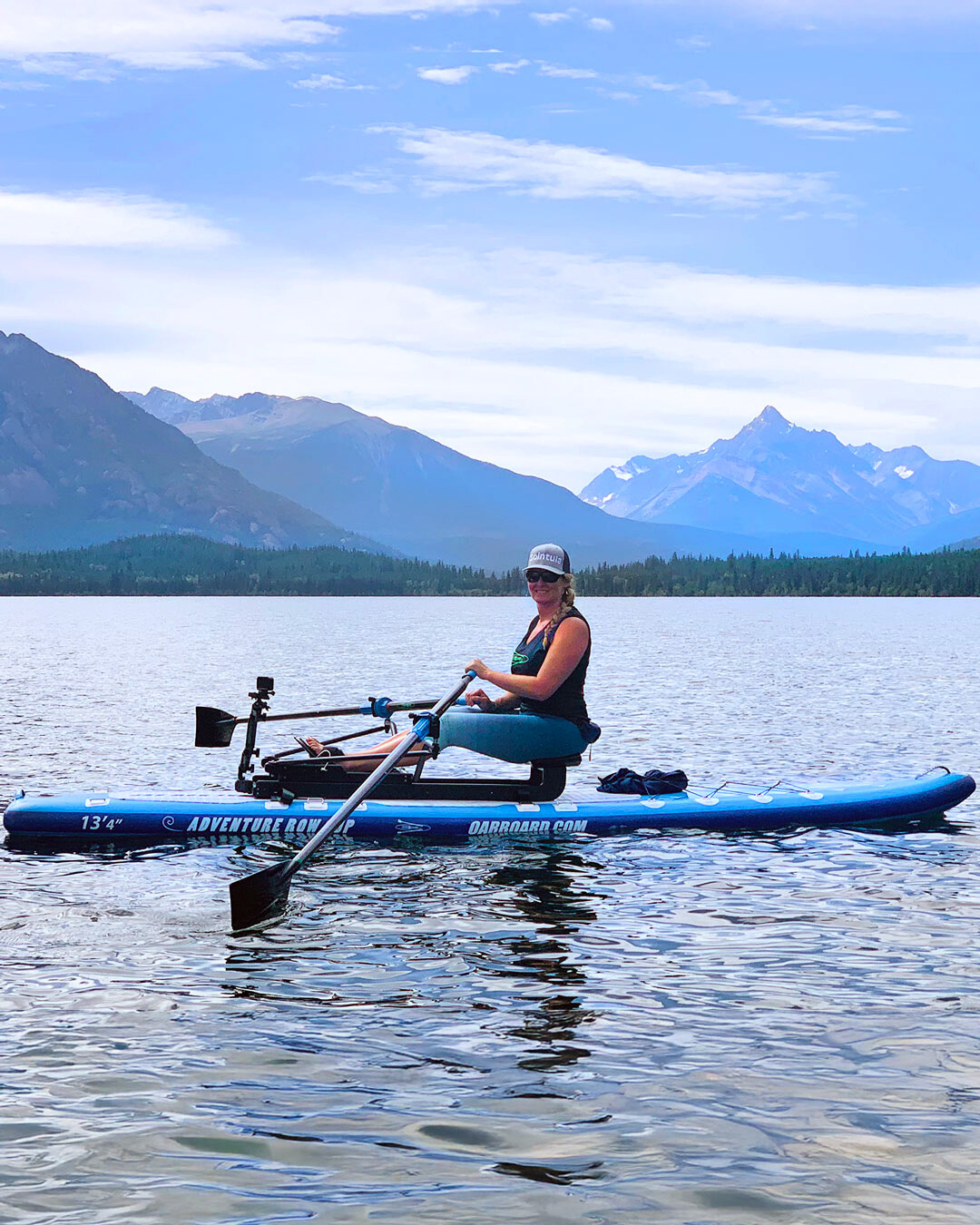 A woman sitting on an Oar Board Rower attached to a paddleboard on a calm lake with mountains in the background under a blue sky.