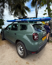 A green SUV parked outdoors on a sandy beach-like surface with palm trees and sky visible. A bright blue paddleboard is mounted on the car roof.