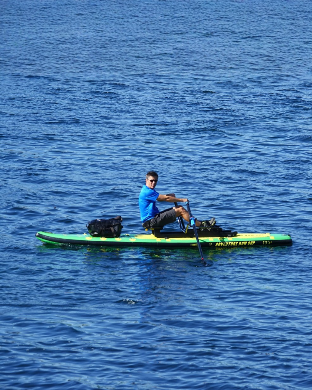 A man in a blue shirt sits rows on a green and yellow stand-up paddleboard on blue water.