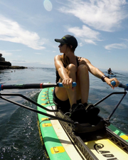 A woman rows a green and yellow stand-up paddleboard on the open sea under a blue sky with white clouds.