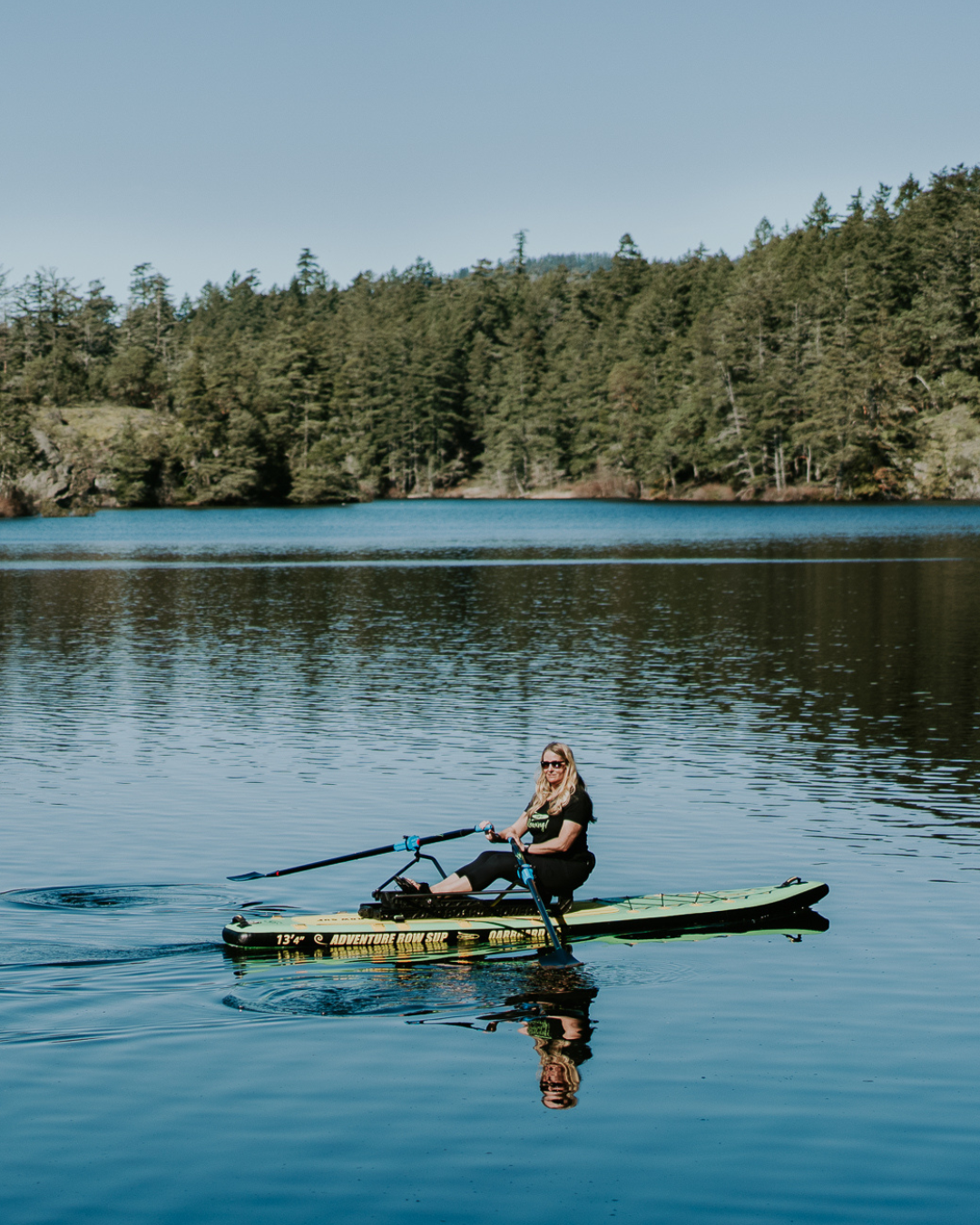 A woman rows a green paddleboard on a calm blue lake with a dense evergreen forest shoreline in the background.