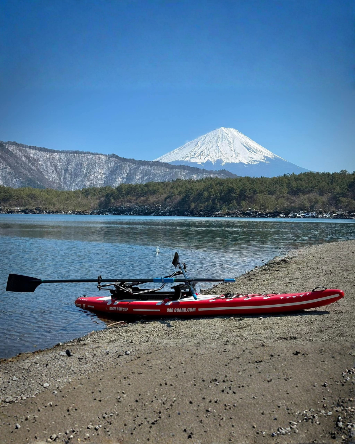 A red paddle board with a black rowing system and black oars attached resting on a sandy shoreline of a lake. With a snowcapped mountain in the background.