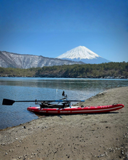A red paddle board with a black rowing system and black oars attached resting on a sandy shoreline of a lake. With a snowcapped mountain in the background.