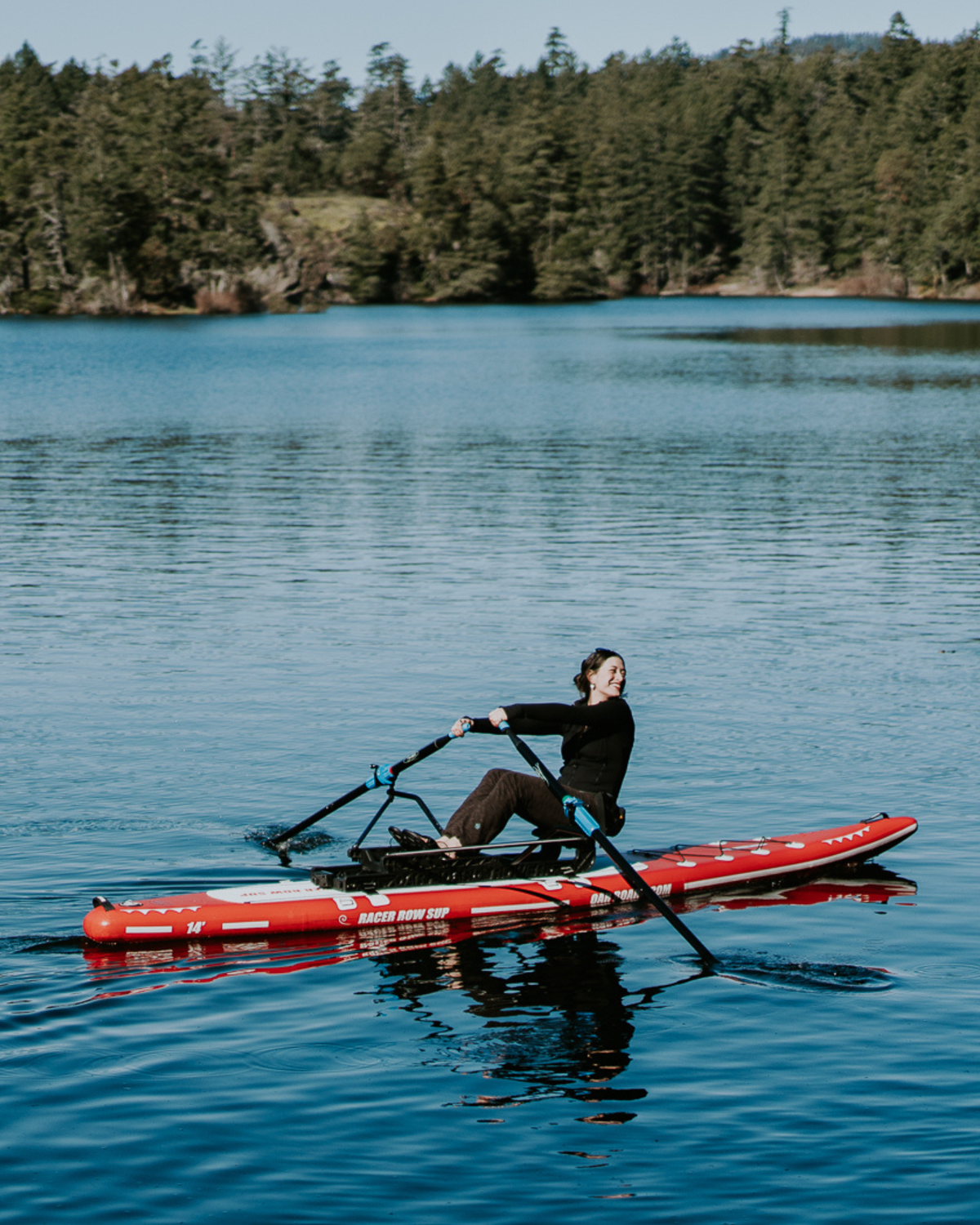 Woman dressed all in black rows a red and white paddle board on a deep blue lake with green tree in the background. 
