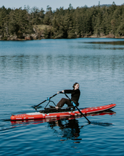 Woman dressed all in black rows a red and white paddle board on a deep blue lake with green tree in the background. 