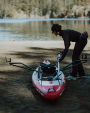 A woman dressed all in black holds a black oar, leaning down to a red and white paddle board that's on a sandy beach of a lake.