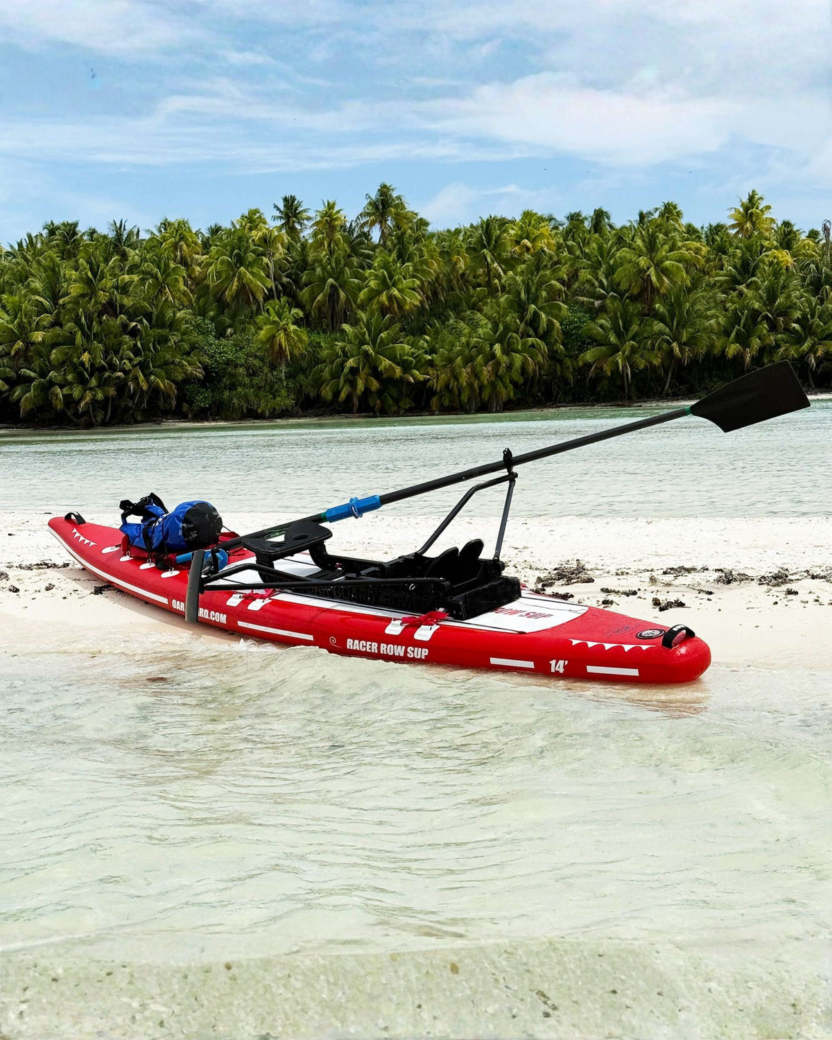 A red paddle board sitting on a white sandy beach with a rowing unit attached and oars in the air. A palm tree lined island is visible in the background.