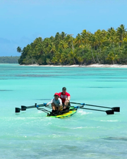 Two people rowing a green paddle board on turquoise water with a tropical palm-lined island in the background.
