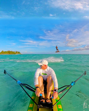 A person in a white long-sleeve shirt and white cap rows a paddleboard in turquoise ocean water under a blue sky, with another person airborne on a board in the distance.