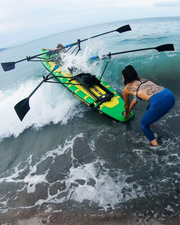 A person in blue leggings and a black bikini stands in shallow ocean water beside a green and yellow stand-up paddleboard with paddles attached, with a wave crashing around it.