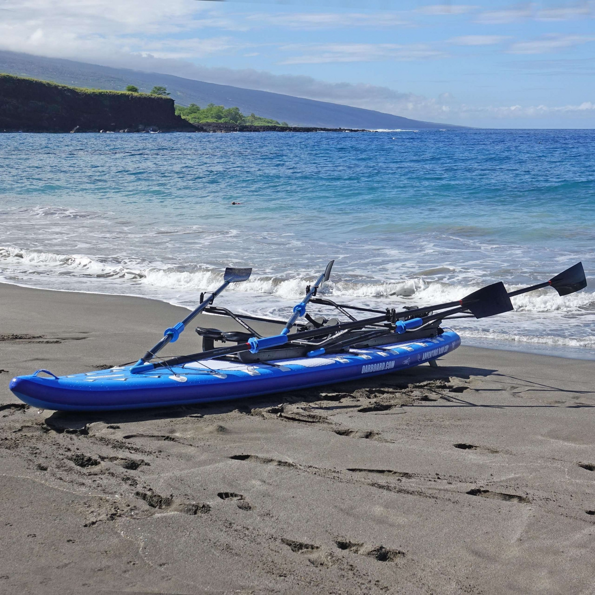 A blue and black dual-person paddle board rests on a sandy beach with the ocean, waves, and distant cliffs in the background.