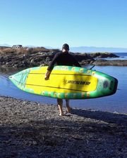 A person carrying a bright yellow and green stand-up paddleboard on a rocky shoreline by the water, photographed from behind under a clear blue sky.