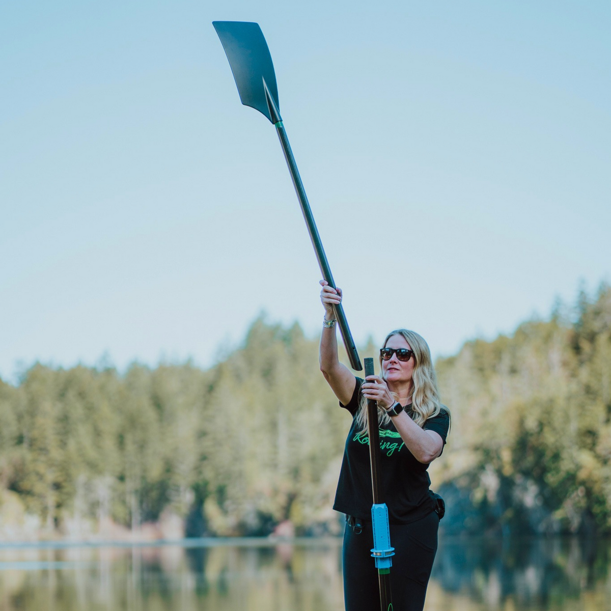 A woman wearing sunglasses and a black t-shirt stands on a lake holding a two-part oar upright, with a forest in the background.