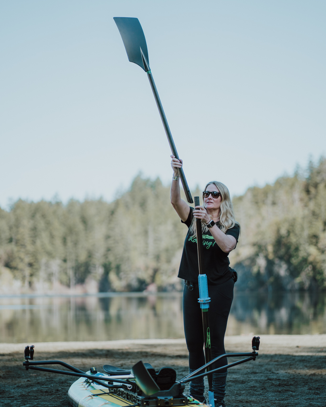 A woman stands outdoors by a lake holding an oar upright, with a paddle and paddle accessories visible in the foreground and trees in the background.