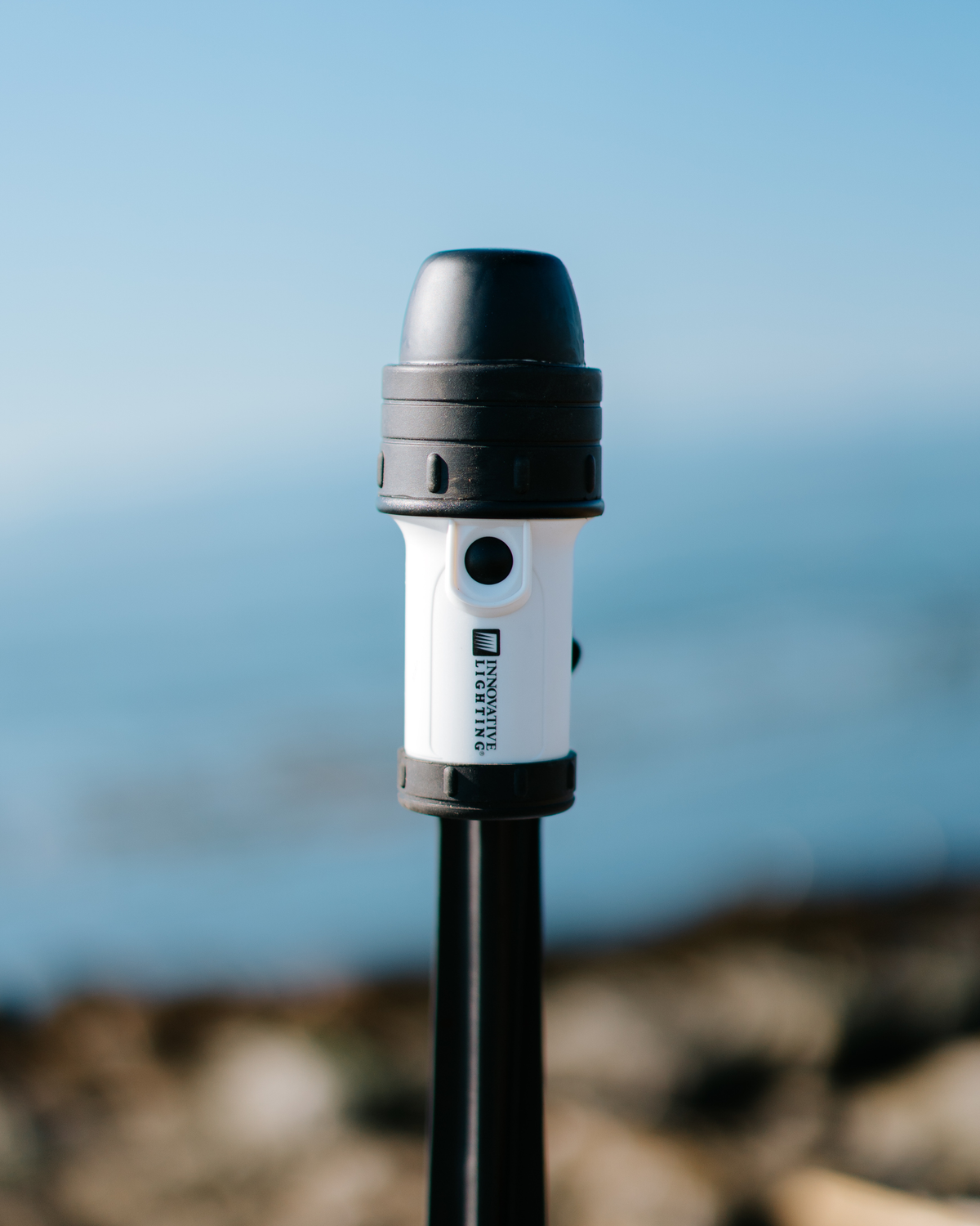 A close-up shot of a marine navigation light mounted on a black pole outdoors, with a blurred coastal background.