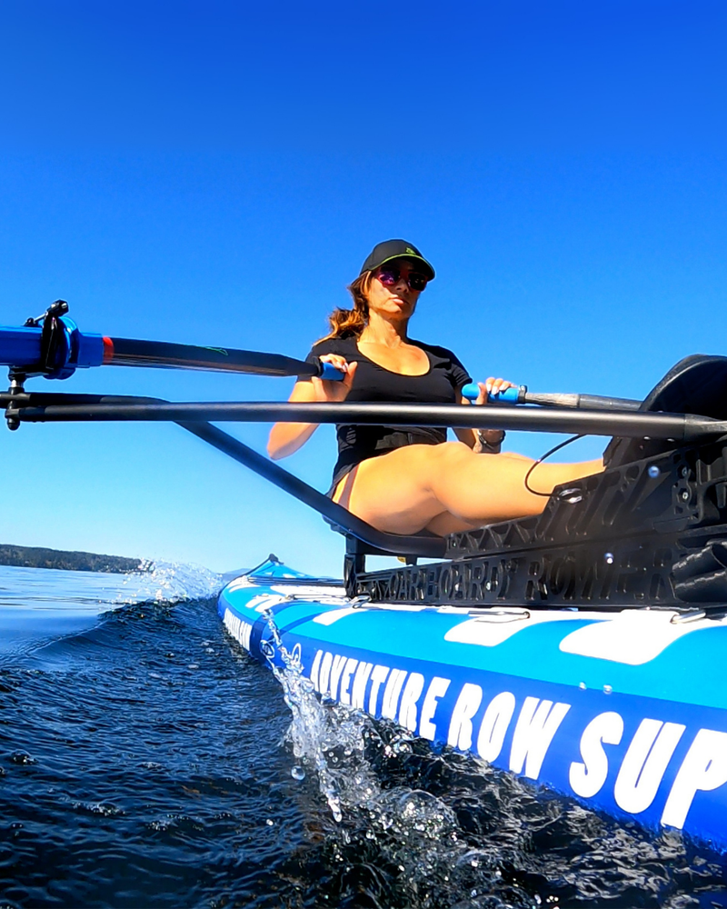 A woman rowing on a blue and white inflatable paddleboard on open water under a clear blue sky.