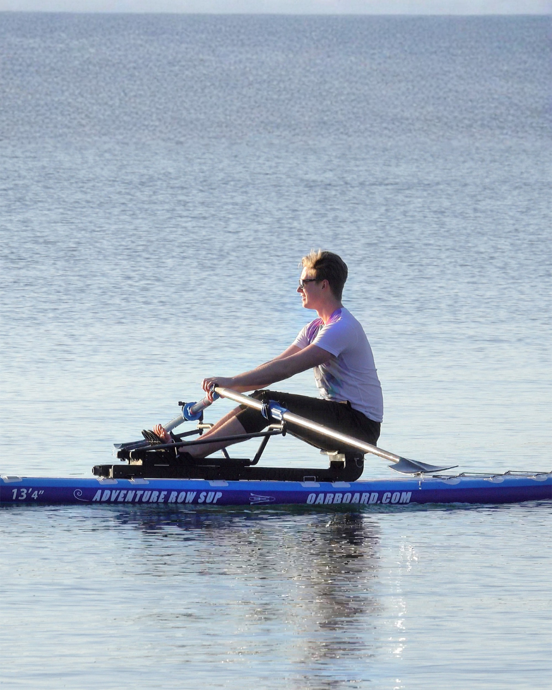 A person rowing on a blue stand-up paddleboard on calm water, wearing a white t-shirt and dark shorts.