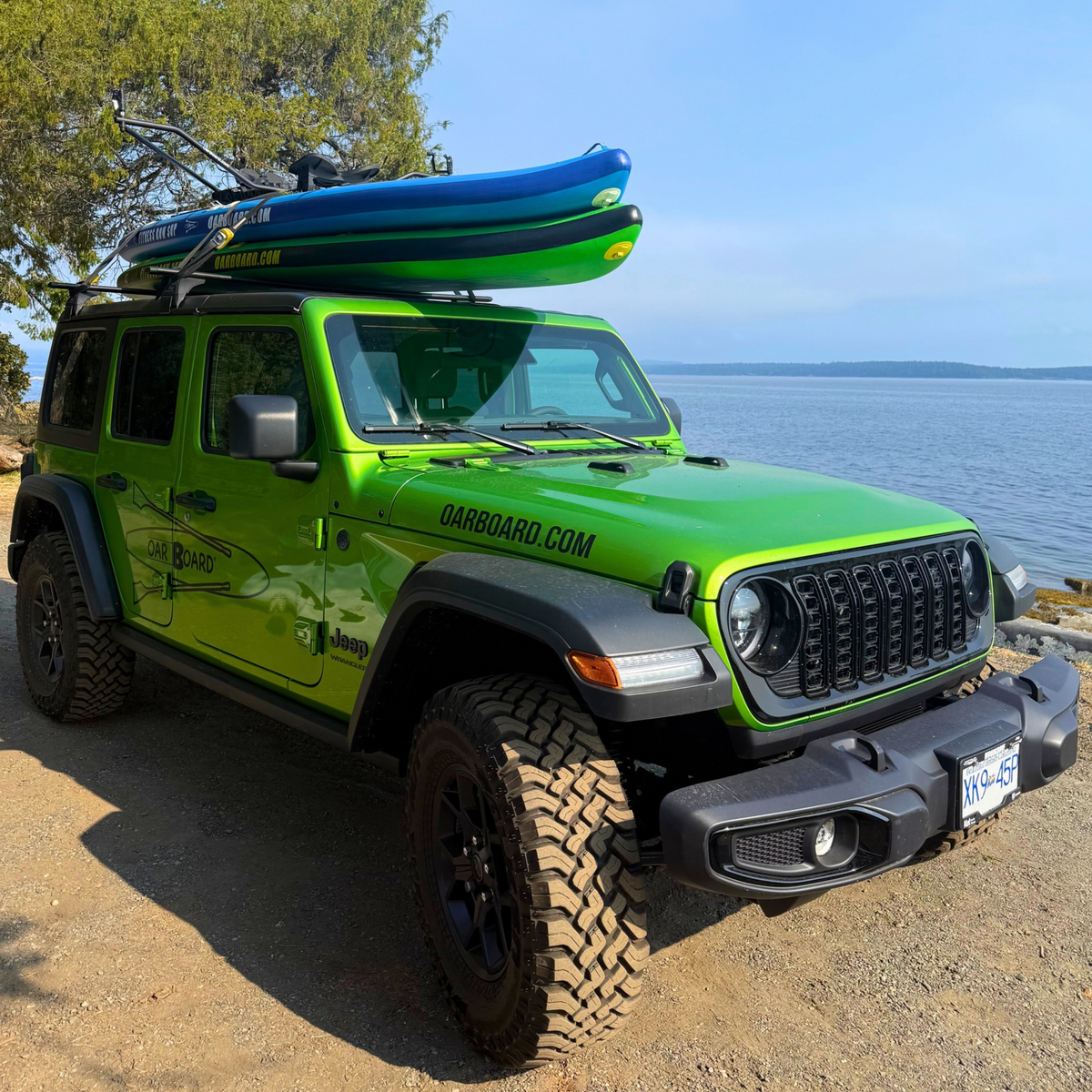 A bright lime green Jeep Wrangler parked outdoors by the water, with a roof-mounted paddle board rack carrying blue and green paddle boards. The vehicle has black off-road tires, a black front bumper, and visible 'OARBOARD.COM'