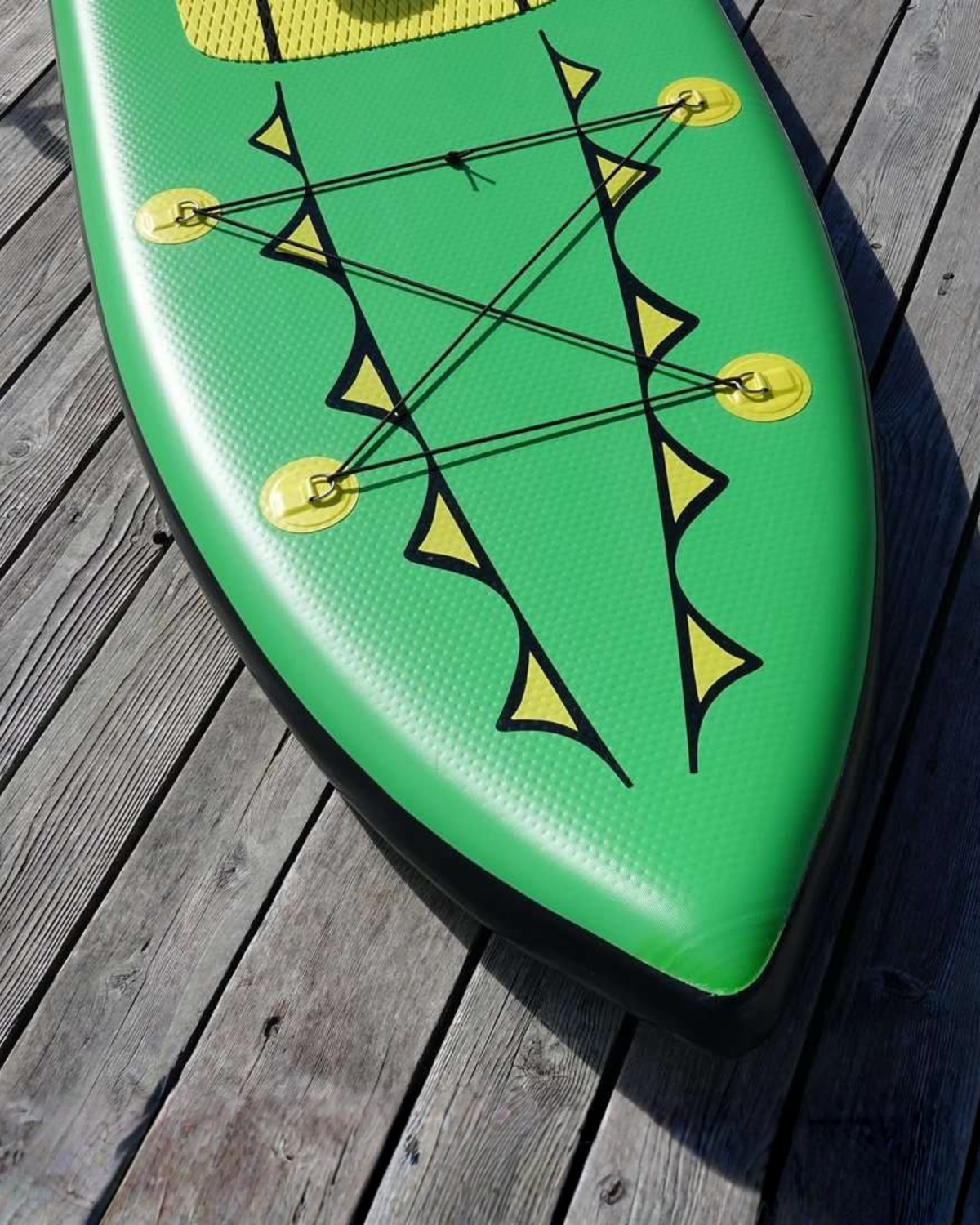A green inflatable stand-up paddle board with yellow and black graphics and bungee cords, photographed on weathered wooden deck planks.
