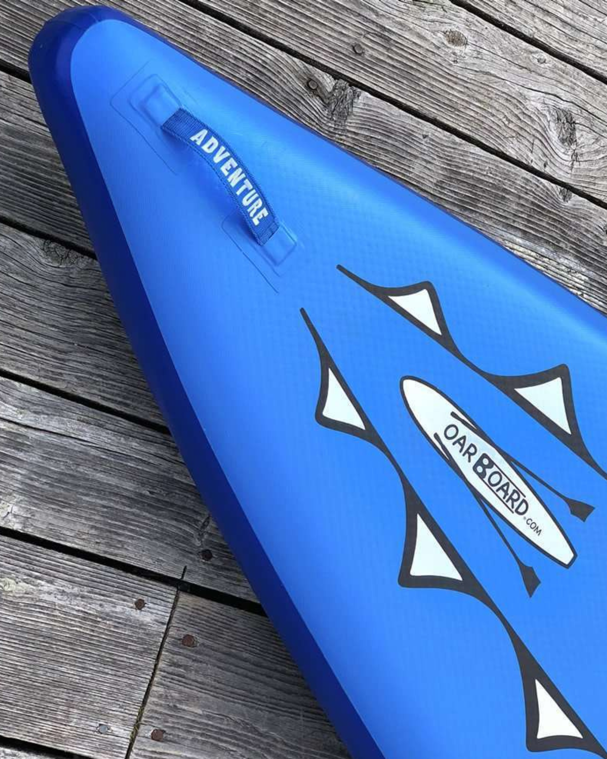 A blue inflatable stand-up paddle board shown diagonally on weathered wooden planks. The board features white and black graphic decals, including a white 'OAR BOARD' logo and a blue 'ADVENTURE' carry handle.