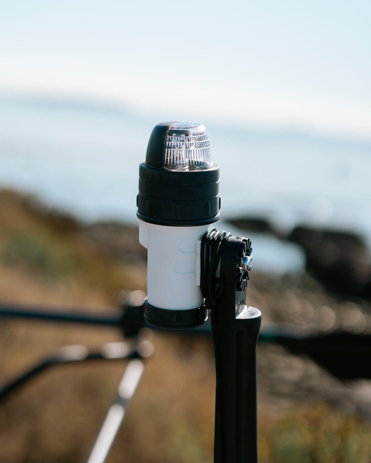 A close-up shot of a marine navigation light mounted on a black pole outdoors, with a blurred coastal background.