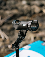 A close-up shot of black fishing rod holder, with a blurred outdoor rocky background and a blue and white surfboard visible at the bottom.