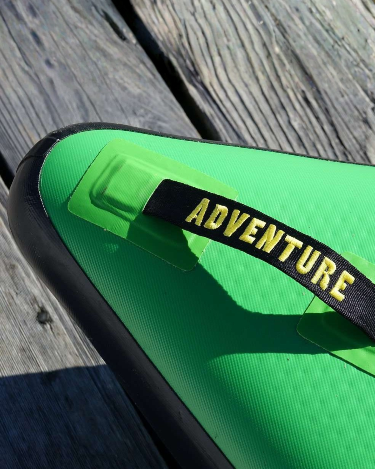 A close-up of a bright green inflatable paddle board with a black trim and strap featuring the word 'ADVENTURE' in yellow lettering, resting on weathered wooden planks.
