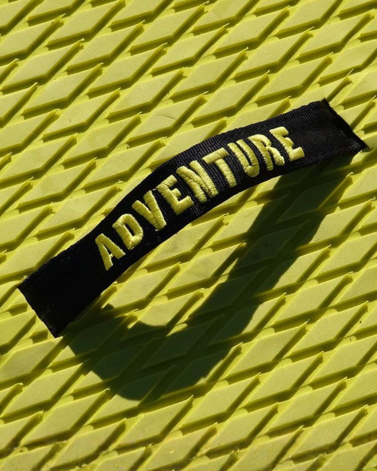 A black fabric patch with the word 'ADVENTURE' embroidered in yellow, placed diagonally on a textured yellow surface.