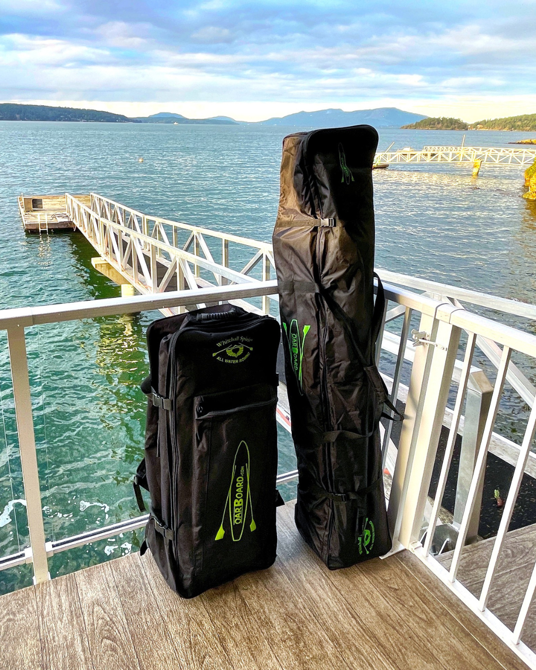 Two black travel bags, one upright and one standing horizontally, placed on a wooden dock with a scenic lake and dock in the background.