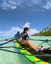 Two people rowing a bright green and yellow double paddle board in clear turquoise tropical water under a blue sky with palm trees in the background.