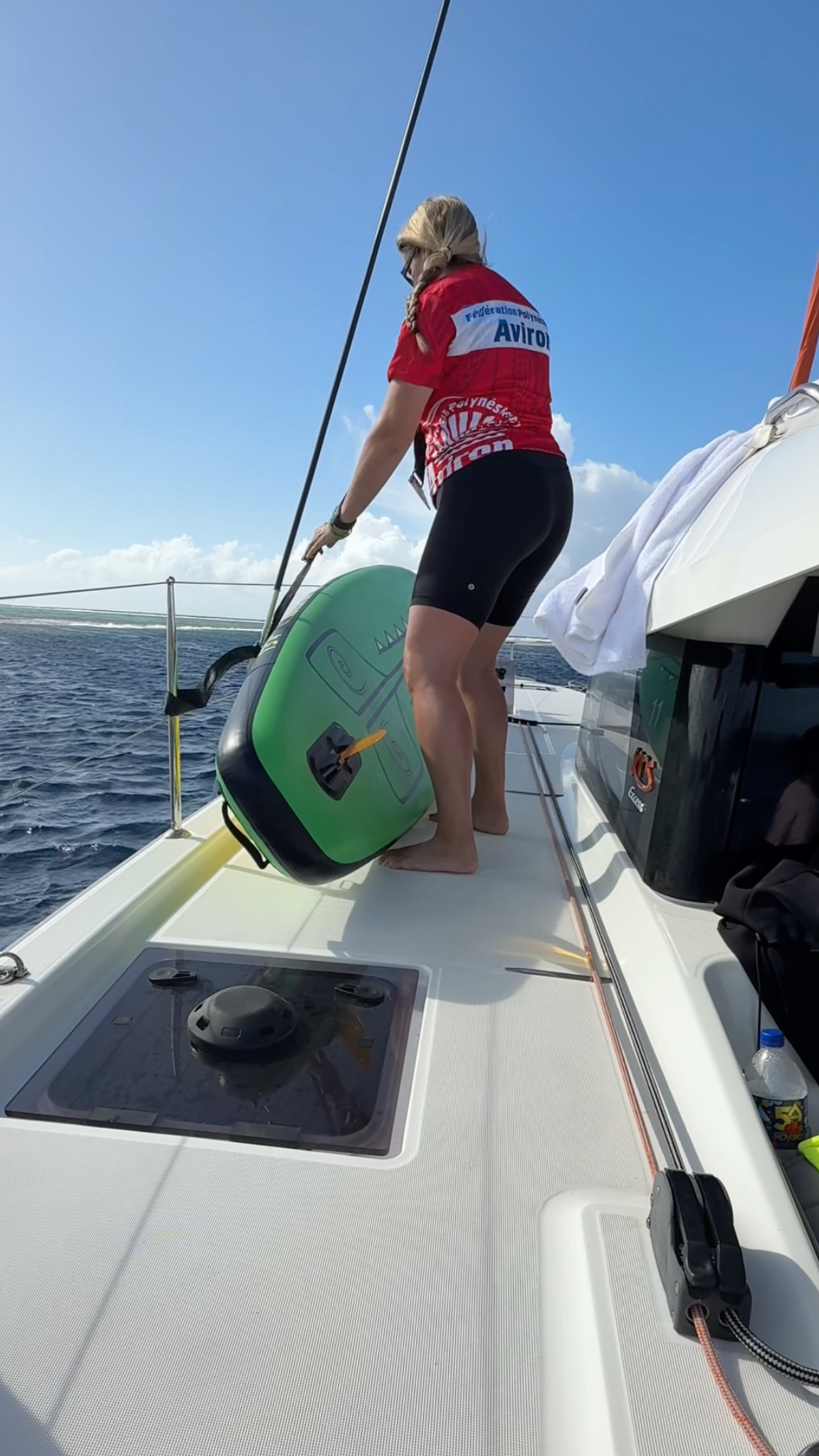 A woman on a sailboat deck wearing a red and white jersey and black shorts, standing next to a green and black paddle board on the deck under a blue sky.