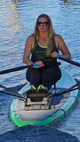 A woman rowing on a paddleboard in open water, wearing a black tank top with green trim and text, black leggings, and sunglasses. The paddleboard is green and white with a textured gray grip pad.