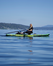 A woman rowing a green and yellow paddleboard on calm blue water, with distant hills and a clear sky in the background.