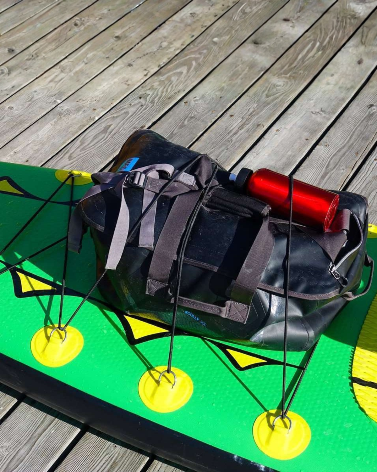 A black waterproof duffel bag with gray straps and a red water bottle, resting on a green paddleboard on a wooden deck.