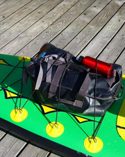 A black waterproof duffel bag with gray straps and a red water bottle, resting on a green paddleboard on a wooden deck.