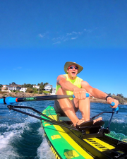 A man wearing a wide brim hat, sunglasses, and a neon yellow shirt is rowing on a green and yellow stand-up paddleboard in blue water under a clear blue sky.
