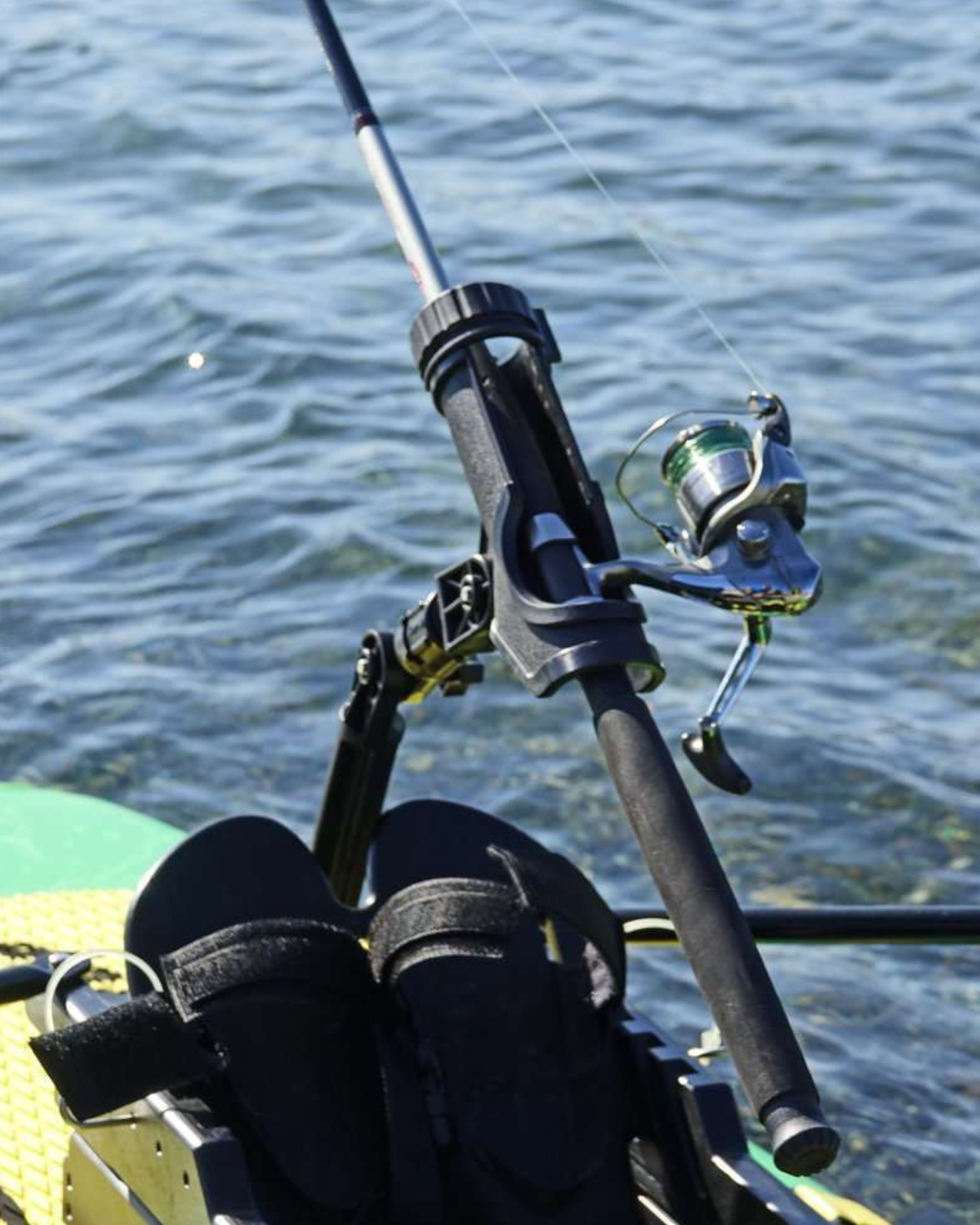 A close-up of a fishing rod mounted on a paddleboard, with a spinning reel attached. The rod and reel are black and silver, set against a background of blue water.