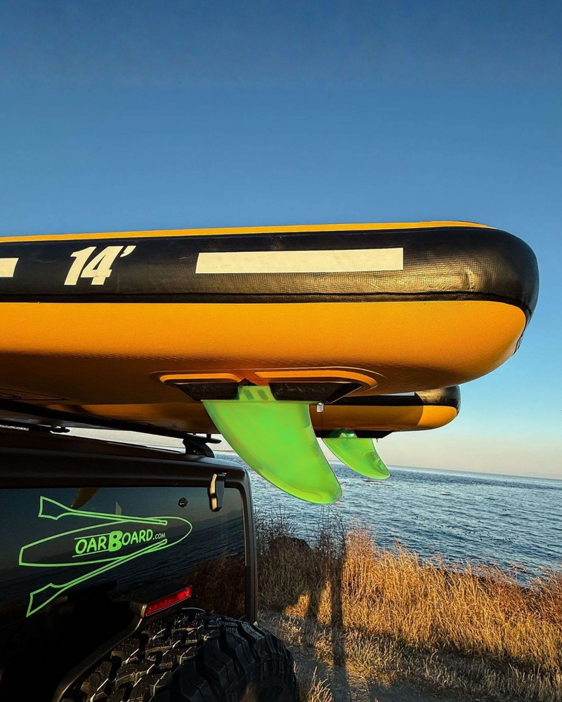 A yellow and black inflatable paddle board mounted on a black vehicle roof rack, with bright green paddle fins visible, photographed outdoors near the water under a blue sky.
