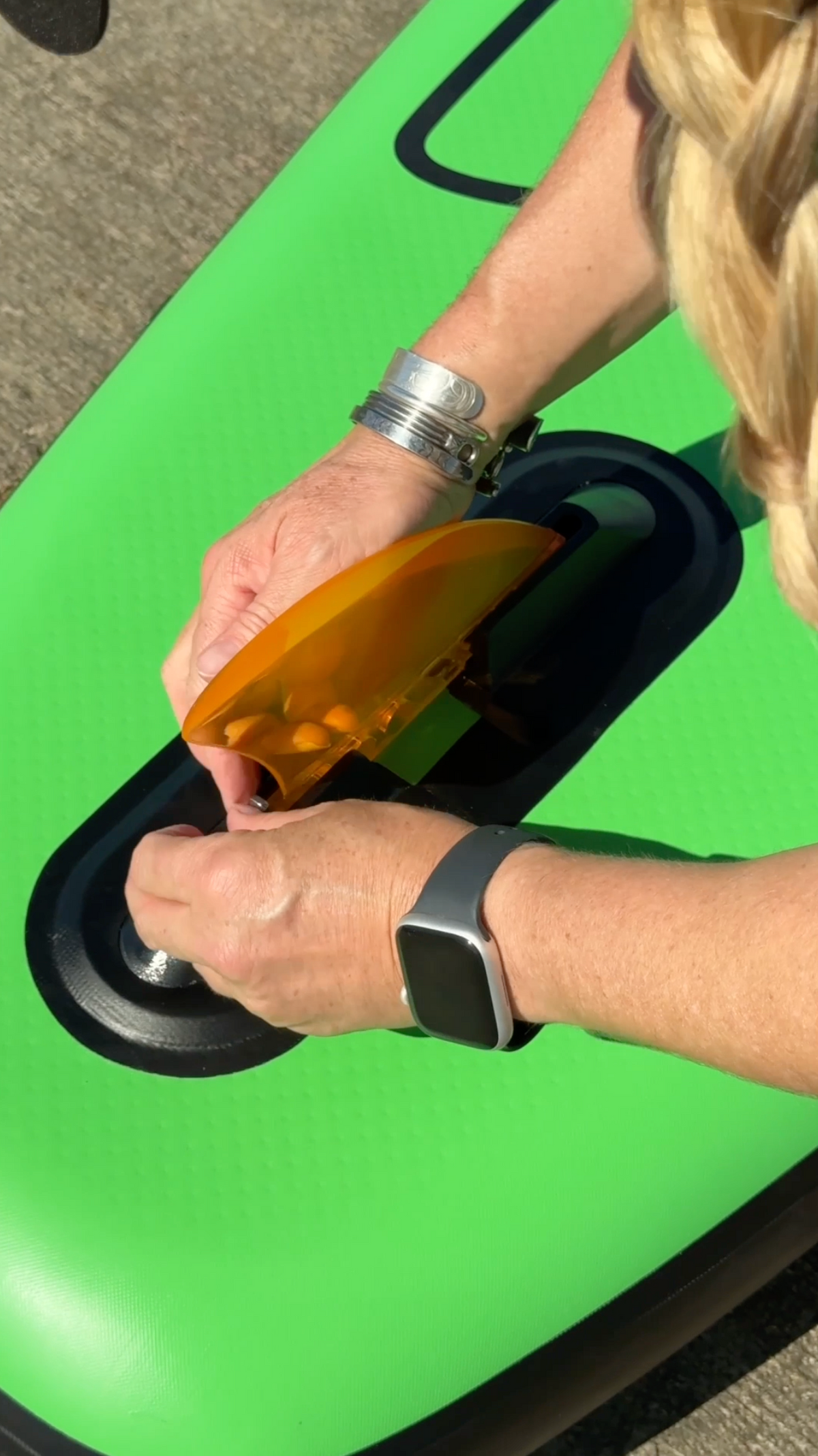A close-up of a person's hands installing an orange translucent paddle board fin on a bright green inflatable paddle board in outdoor sunlight.