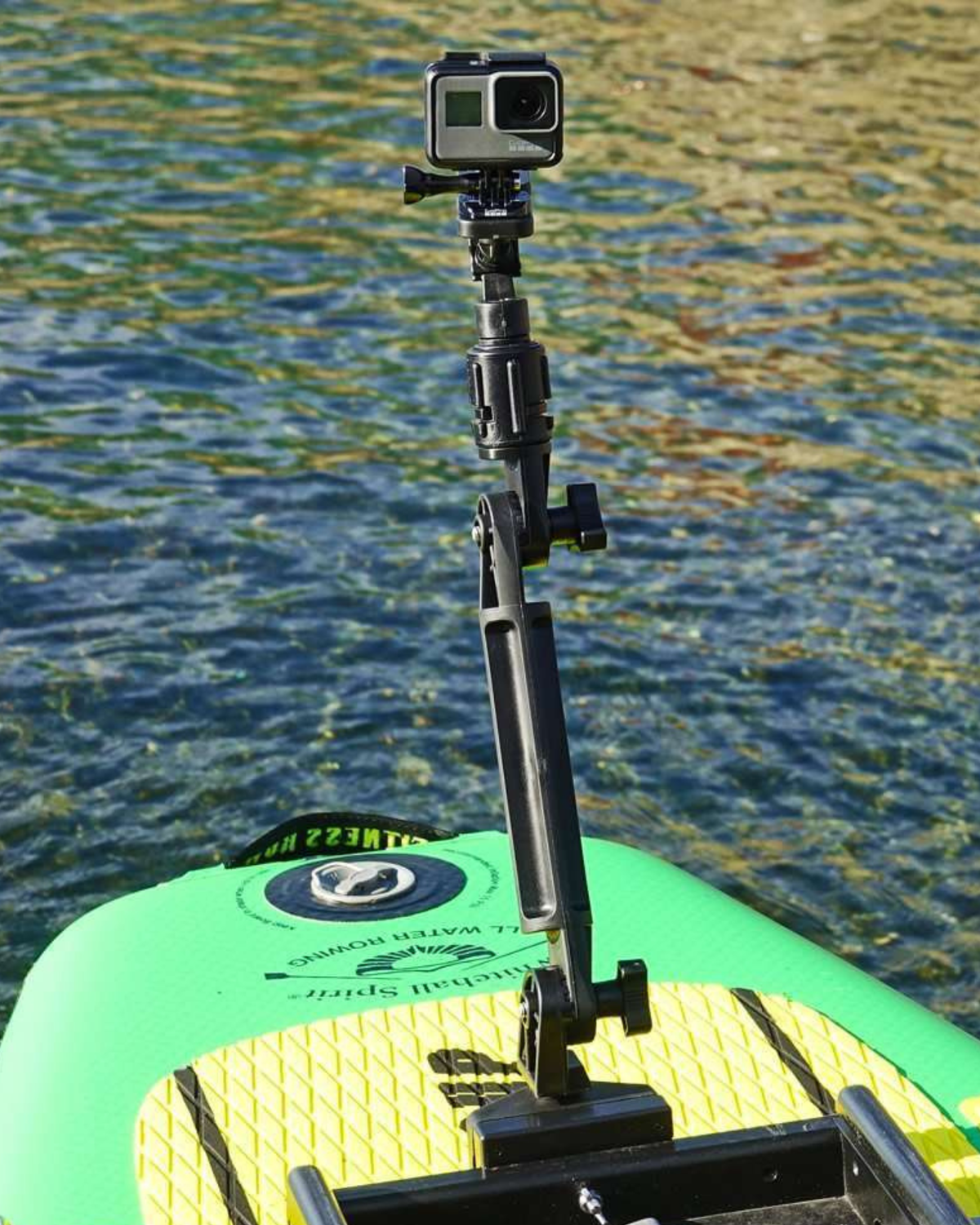 A black adjustable mounting pole with a mounted GoPro camera, shown attached to the front of a green and yellow paddle board on water.
