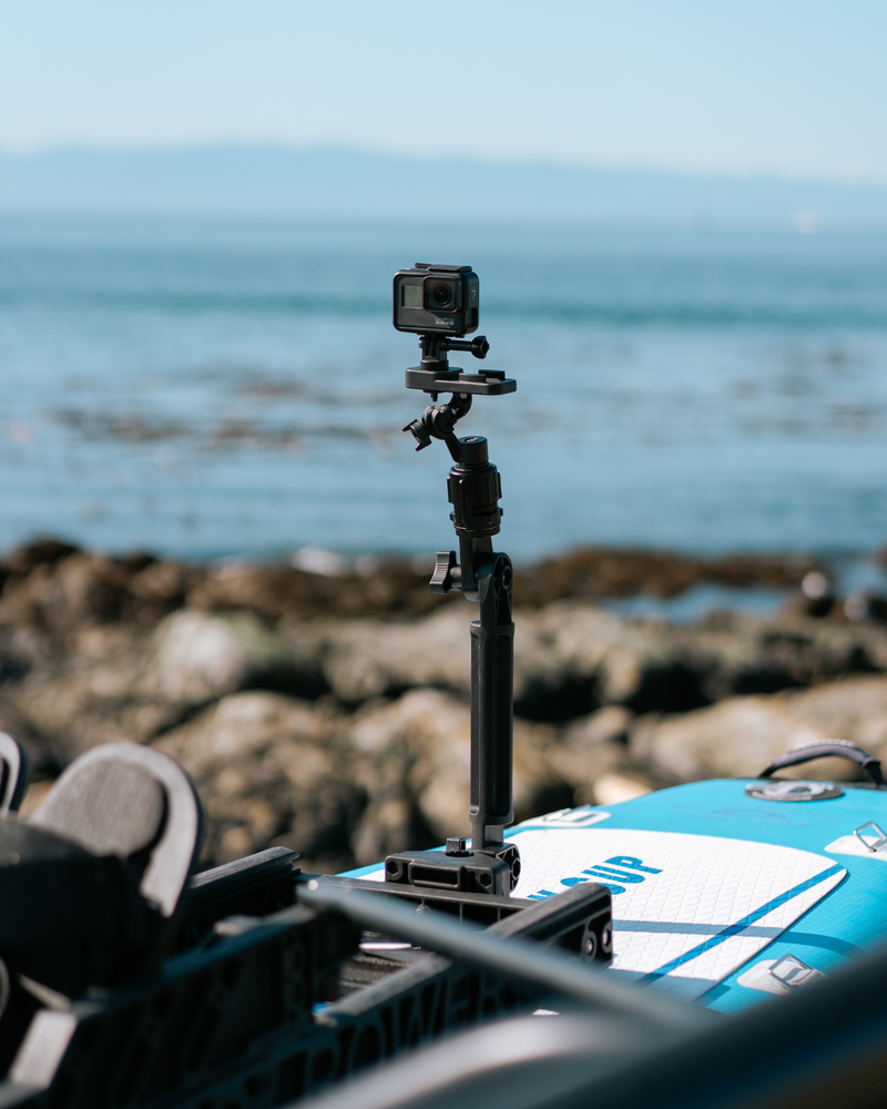 A black action camera mounted on a vertical tripod stand attached to a blue paddleboard on a rocky shoreline with the ocean in the background.