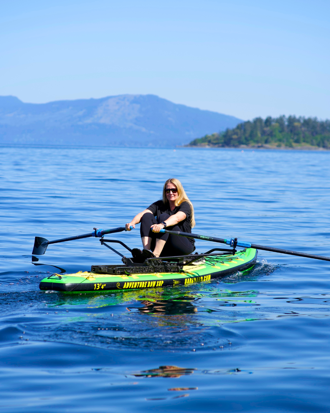 A woman rows on a green and black stand-up paddleboard on calm blue water.