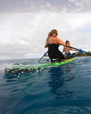 Two women rowing a green and black paddle board on deep blue ocean water under a cloudy sky.