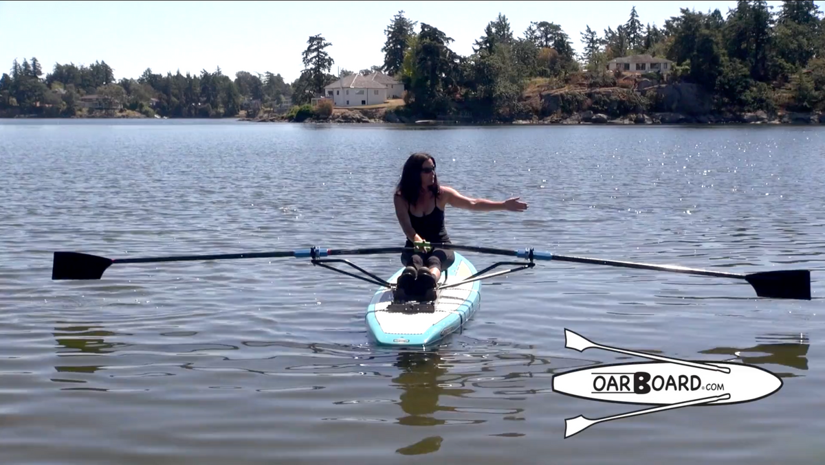 A woman rowing on a stand-up paddleboard on a calm lake, with trees and houses visible in the background. The paddle extends horizontally on both sides, and an 'OAR BOARD'