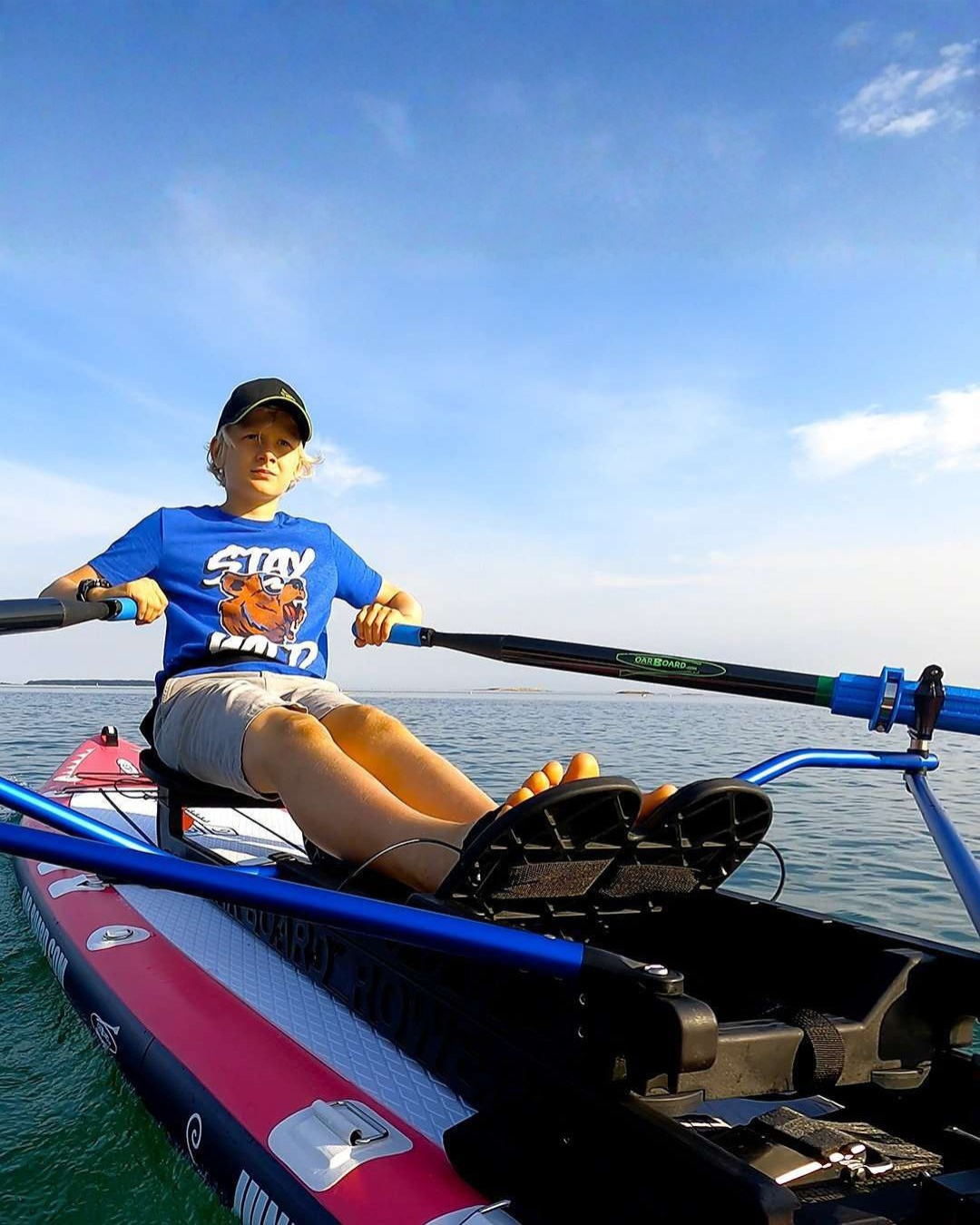 A person rowing the junior rowing kit on a pink inflatable stand-up paddle board on open water under a blue sky.