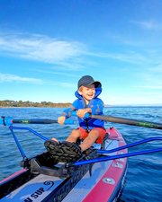 A young boy wearing a blue life jacket and a dark cap is smiling while rowing in a pink and blue paddle board. 