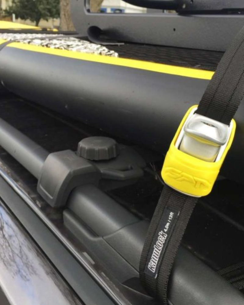 Close-up of a black roof rack strap with a bright yellow quick-release buckle, attached to a black roof bar on a vehicle.