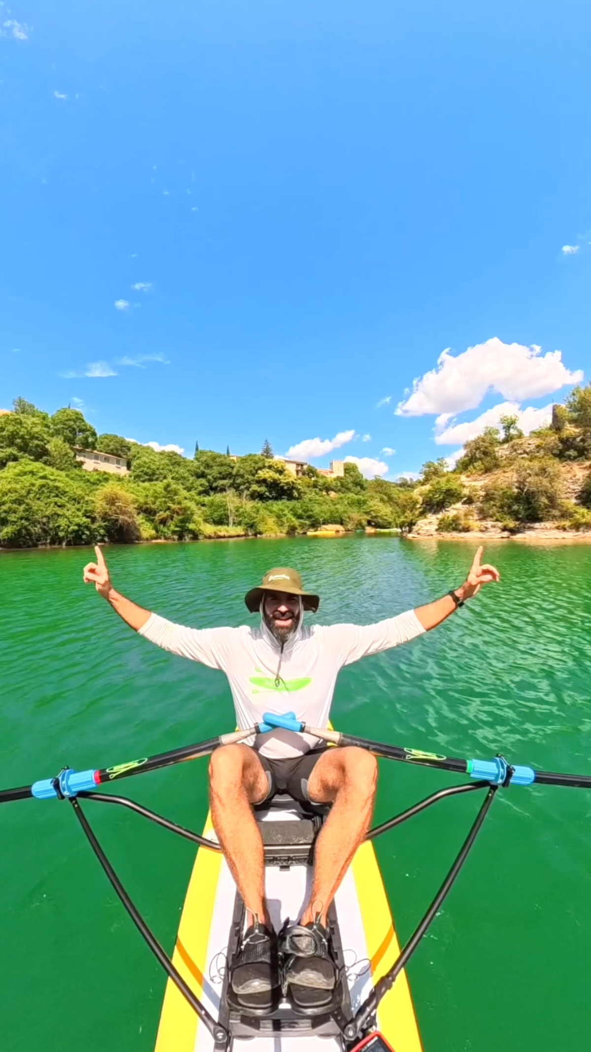 A man smiling sitting on a yellow and white paddle board with his arms in the air pointing his index finger up to the sky. 