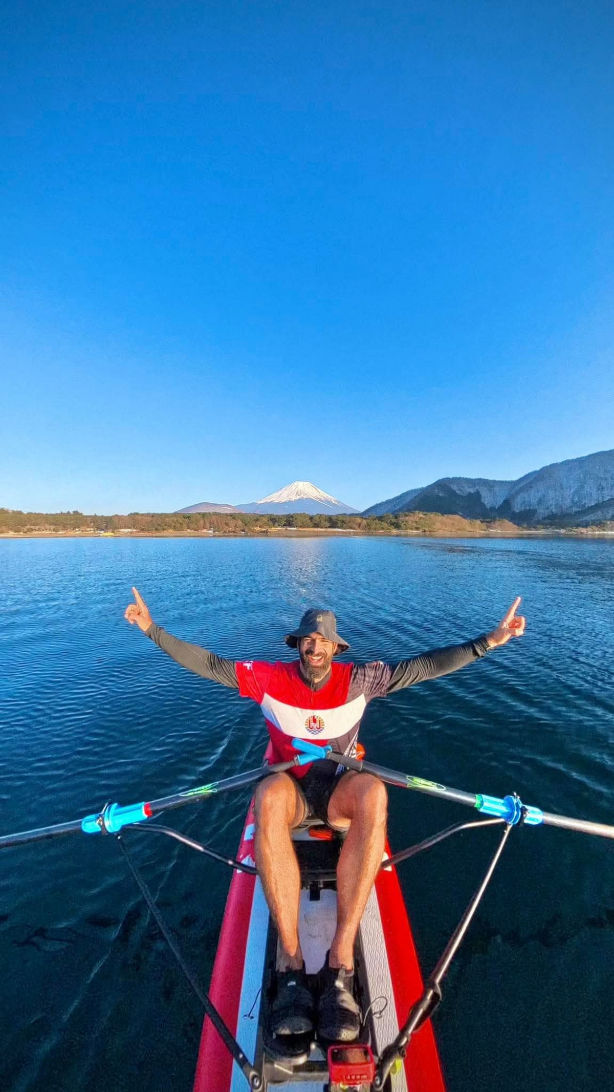 A man wearing a red, white, and blue athletic jersey sits in the bow of a paddleboard on a calm lake, with mountains and a snow-capped peak in the background under a clear blue sky.