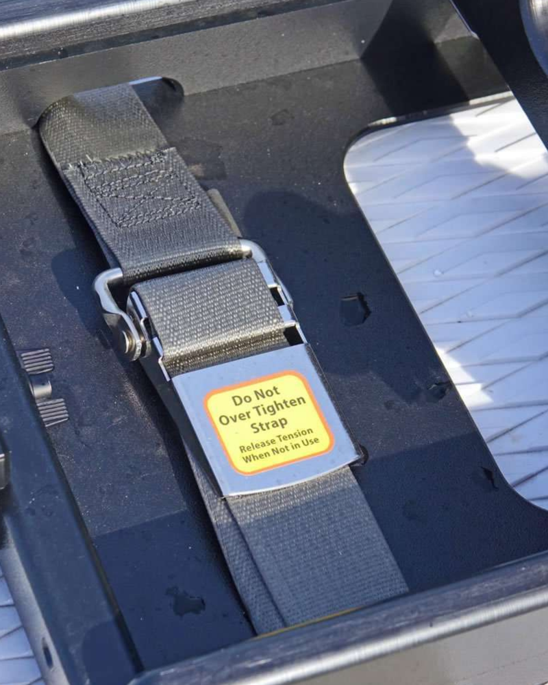 A close-up of a black strap with a silver buckle and a yellow warning label that reads 'Do Not Over Tighten Strap' and 'Release Strap When Not in Use,' shown inside a dark compartment.